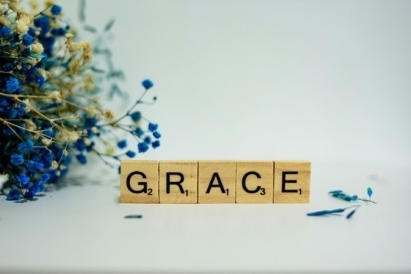 Scrabble tiles that spell out grace on a white background with blue flowers on the left side. 